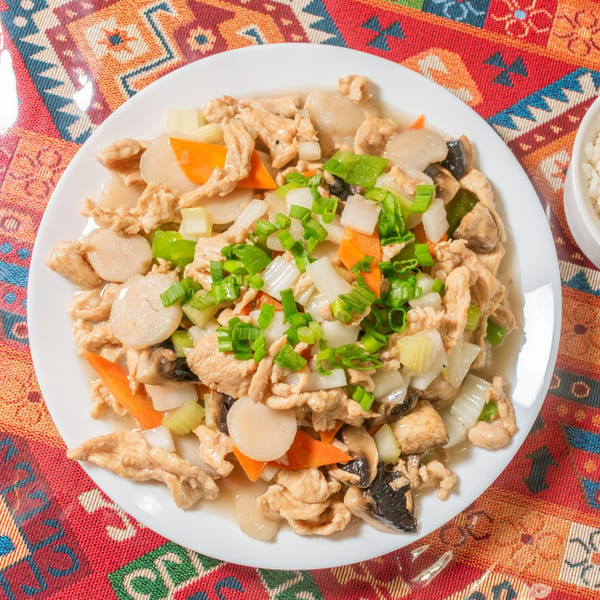 A white plate of stir-fried chicken with vegetables, including carrots, onions, and mushrooms, on a colorful tablecloth.