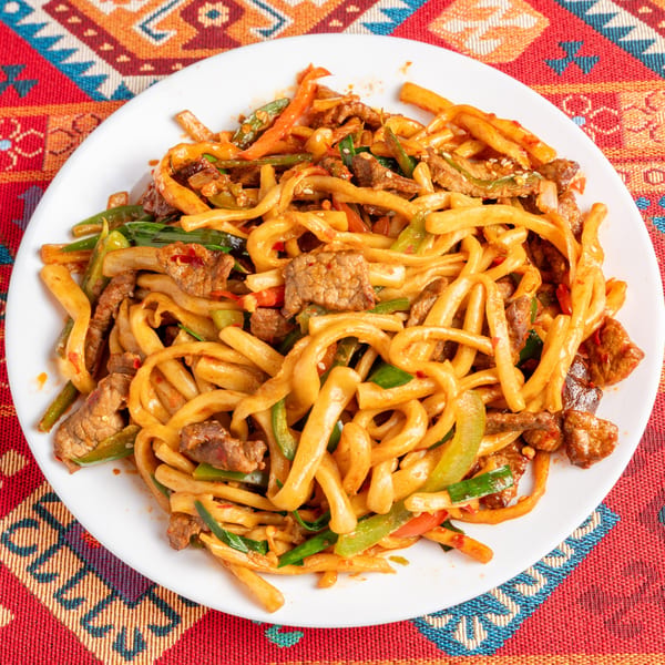 A plate of stir-fried noodles with beef, green peppers, and onions on a colorful patterned tablecloth.