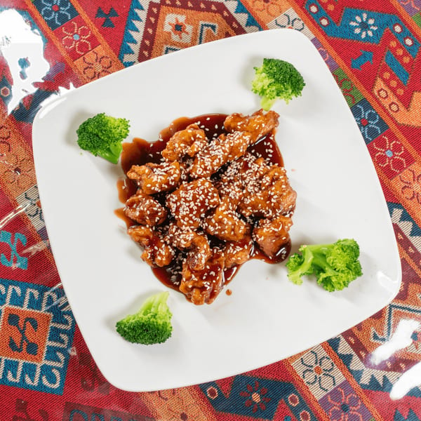 Sesame chicken with broccoli florets on a white plate, set on a colorful patterned tablecloth.