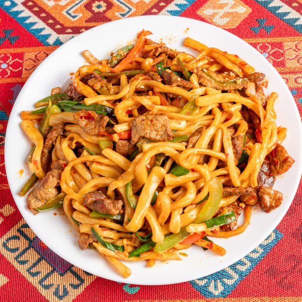 A white plate of stir-fried noodles with beef, green peppers, and onions on a colorful patterned tablecloth.