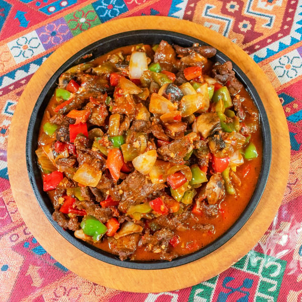 Sizzling beef and vegetable stir-fry in a black skillet on a wooden board, atop a colorful patterned tablecloth.