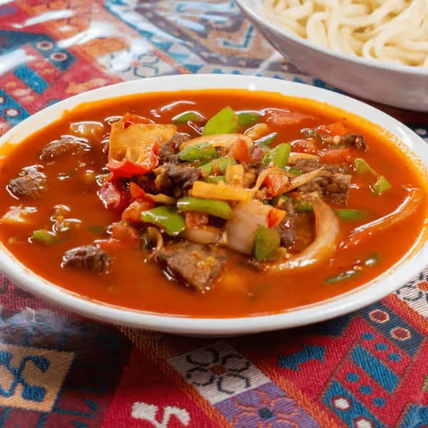 A bowl of vibrant red stew with beef, green peppers, and onions, next to noodles.