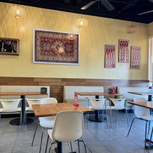 Interior of Turan Uyghur Kitchen with yellow walls, wooden tables, white chairs, and patterned wall art.