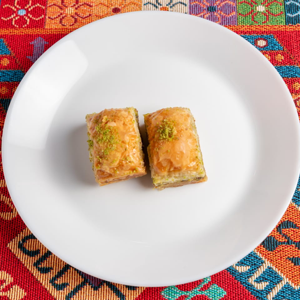 Two pieces of baklava with pistachio sprinkles on a white plate, set on a colorful patterned tablecloth.