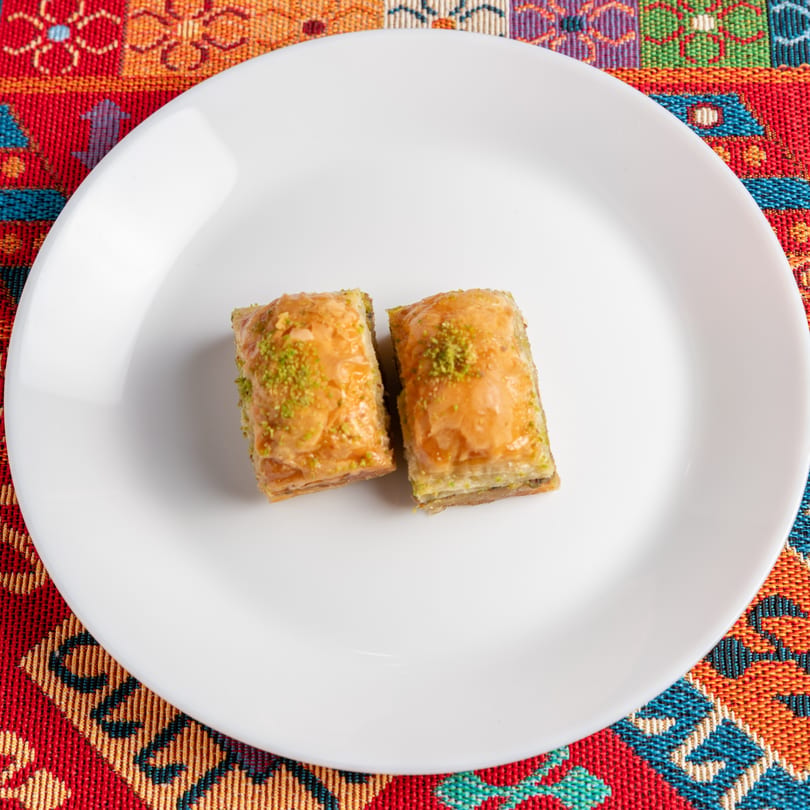 Two pieces of baklava with pistachio sprinkles on a white plate, set on a colorful patterned tablecloth.