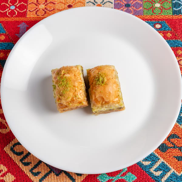 Two pieces of baklava with chopped pistachios on a white plate, on a colorful patterned tablecloth.