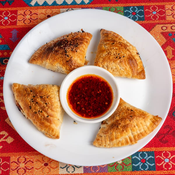 Four golden-brown pastries with sesame seeds, arranged around a small bowl of red chili sauce on a white plate.