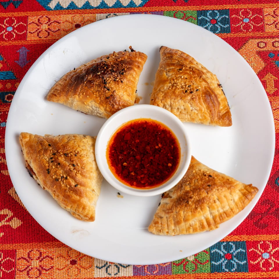 Four golden-brown pastries with sesame seeds, arranged around a small bowl of red chili sauce on a white plate.