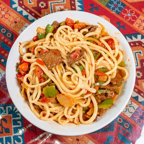 A white plate of stir-fried noodles with beef, green peppers, and tomatoes on a colorful patterned tablecloth.