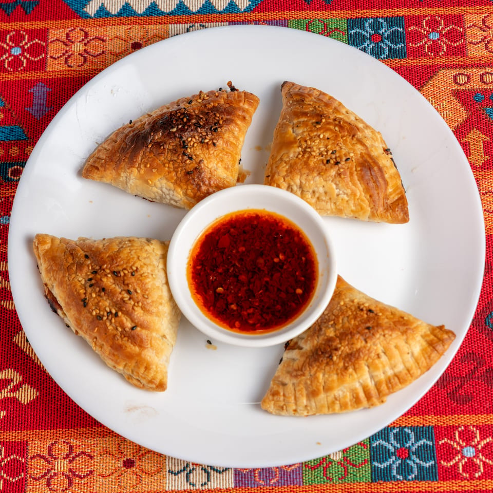 Four golden-brown pastries with sesame seeds surround a small bowl of red chili oil on a white plate.