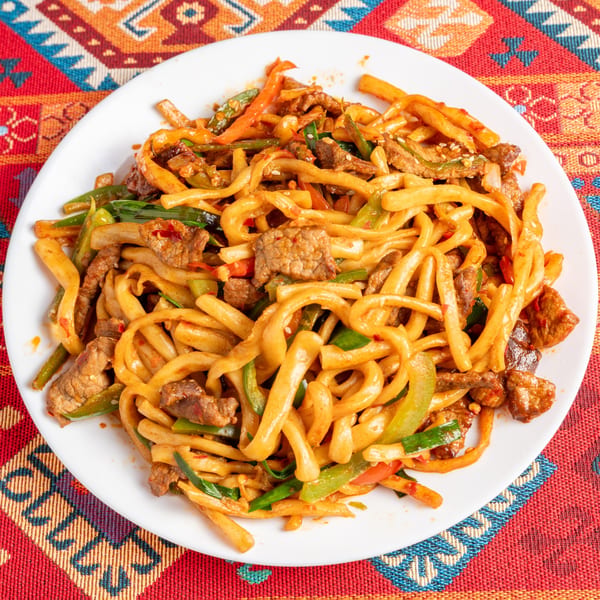 A white plate of stir-fried noodles with beef, green onions, and bell peppers on a colorful patterned tablecloth.