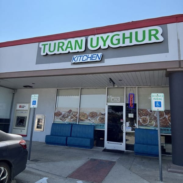 Exterior of Turan Uyghur Kitchen with a large sign, blue benches, and food posters in windows.