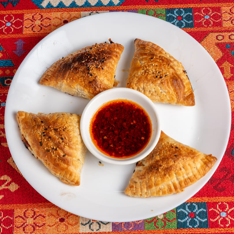 Four golden-brown pastries with sesame seeds surround a small bowl of red chili oil on a white plate.