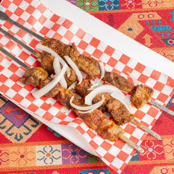 Four skewers of grilled meat with sliced onions on red and white checkered paper, on a colorful patterned tablecloth.