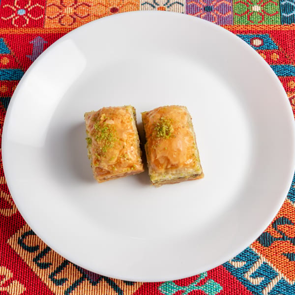 Two pieces of baklava with pistachio sprinkles on a white plate, set on a colorful patterned tablecloth.