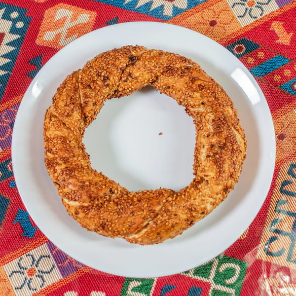 A sesame-covered simit, a Turkish bagel, on a white plate over a colorful patterned tablecloth.
