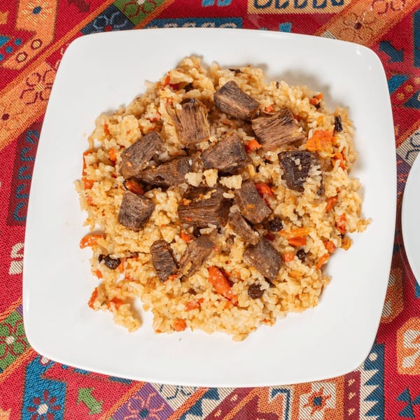 A white square plate of pilaf with chunks of beef, carrots, and raisins on a colorful patterned tablecloth.