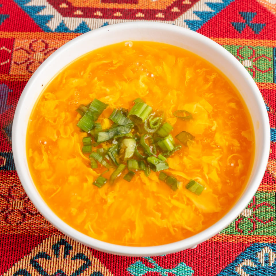 A white bowl of bright orange egg drop soup, garnished with chopped green onions, on a colorful patterned tablecloth.