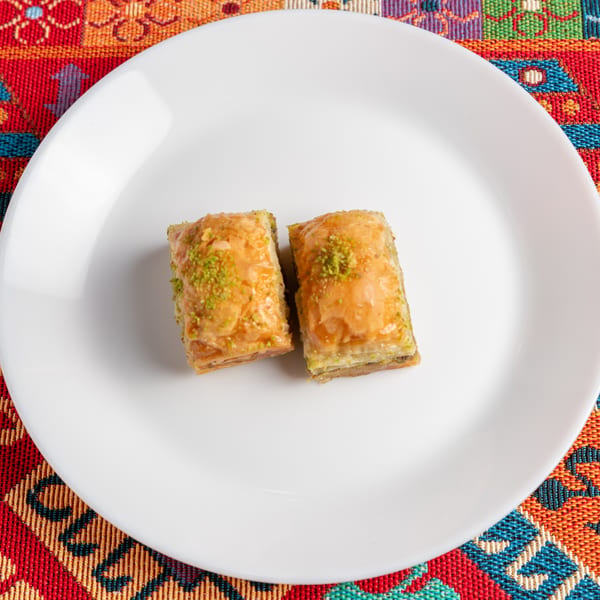 Two pieces of baklava with chopped pistachios on a white plate, on a colorful patterned tablecloth.