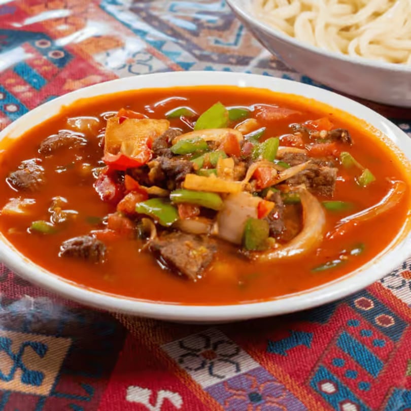 A bowl of vibrant red stew with beef, green peppers, and onions, next to noodles.