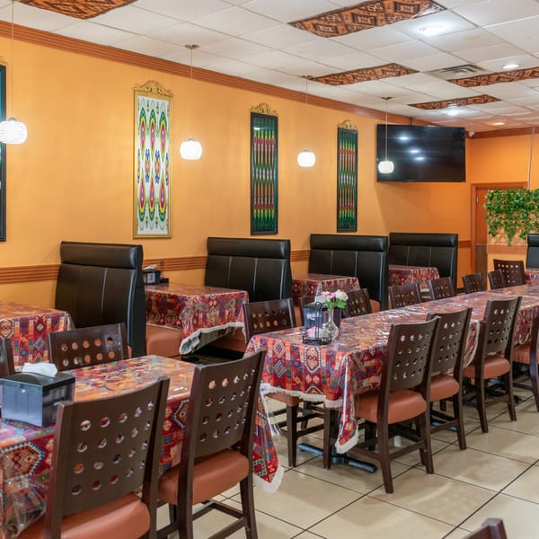 Restaurant interior with orange walls, patterned tablecloths, dark chairs, and booths, under decorative lighting.
