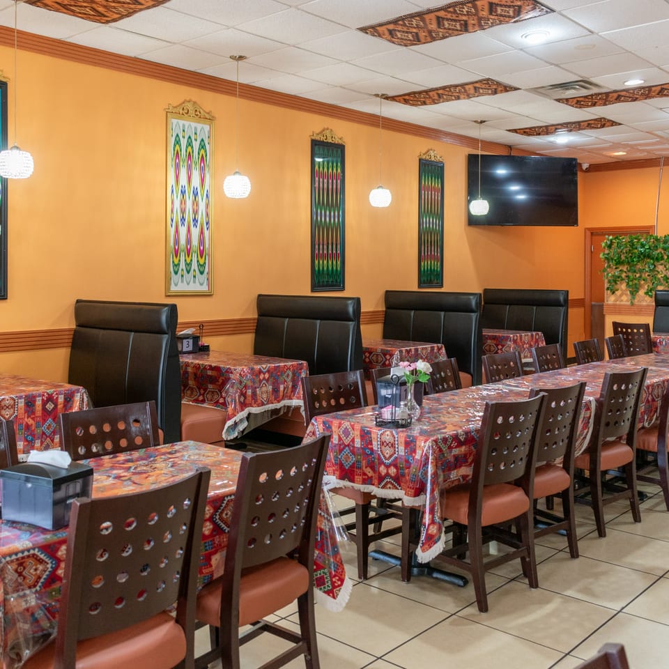 Restaurant interior with orange walls, patterned tablecloths, dark chairs, and booths, under decorative lighting.
