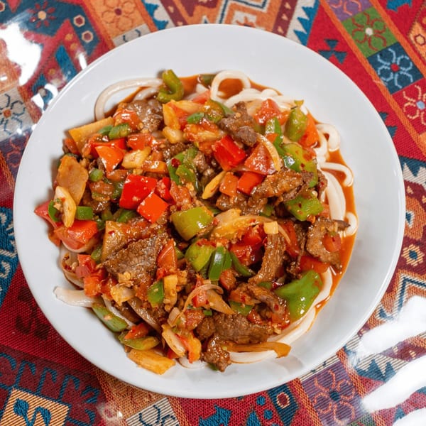 A white plate of laghman noodles topped with beef, red and green bell peppers, and onions.