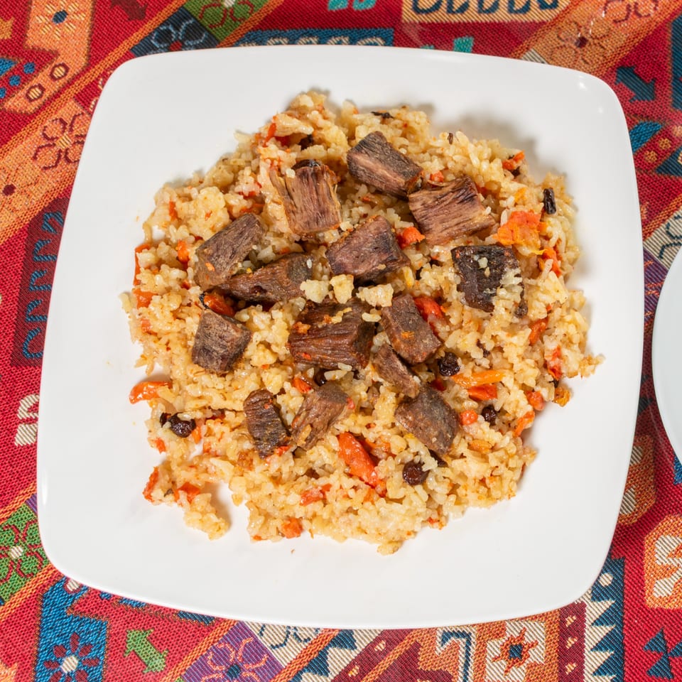 A white square plate filled with pilaf, featuring rice, carrots, raisins, and chunks of beef.
