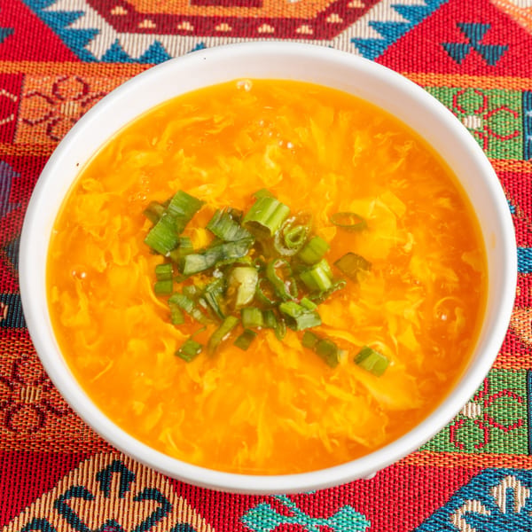 Egg drop soup in a white bowl, garnished with green onions, on a colorful patterned placemat.