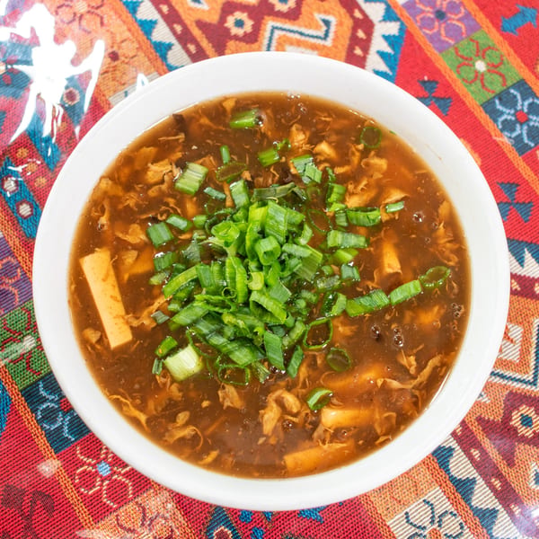 Hot and sour soup with tofu, egg, and green onions in a white bowl on a colorful tablecloth.